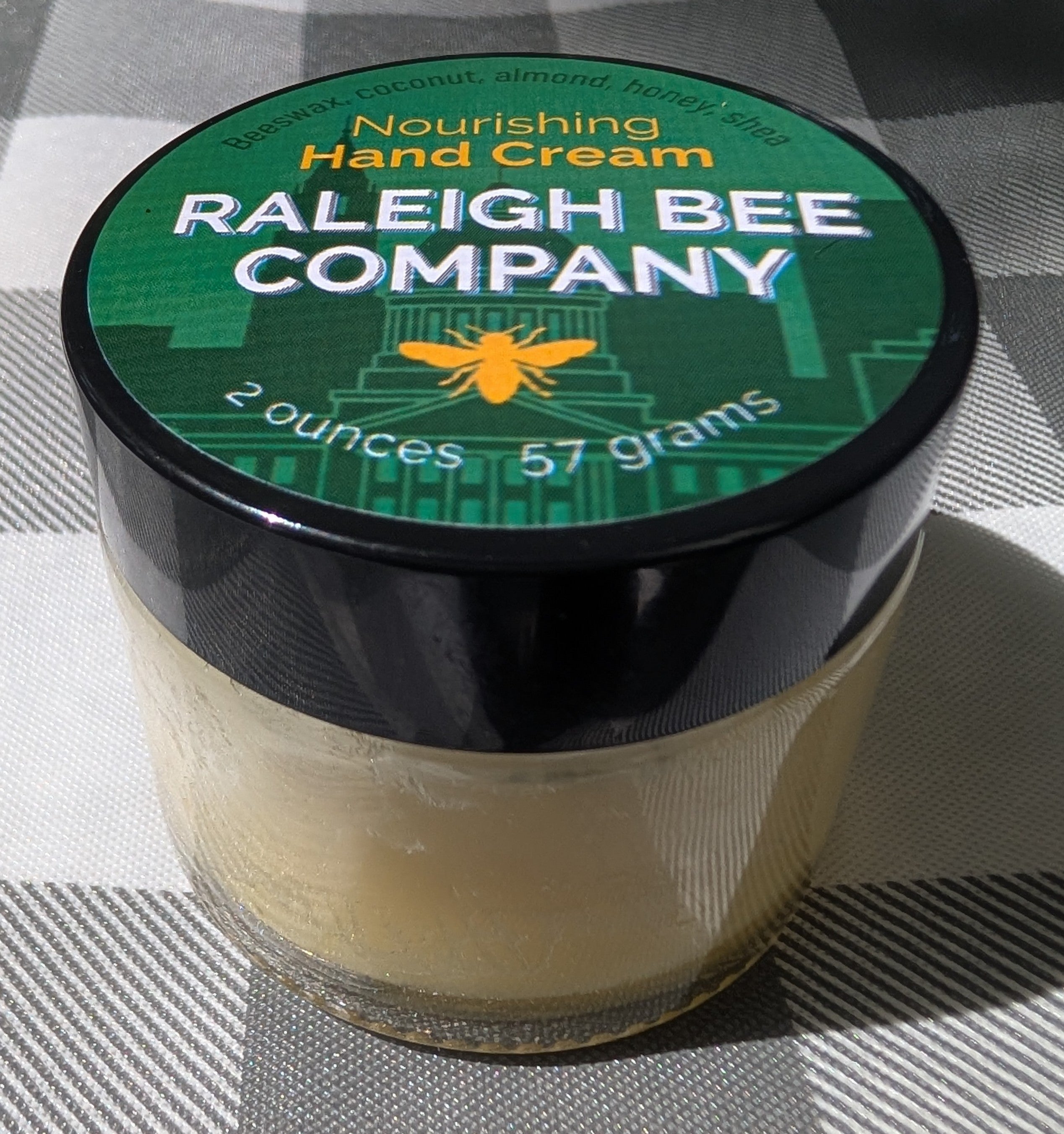 Jar of Raleigh Bee Company hand cream on a checkered fabric background. Contains: Beeswax, coconut oil, almond oil, honey, and shea butter.