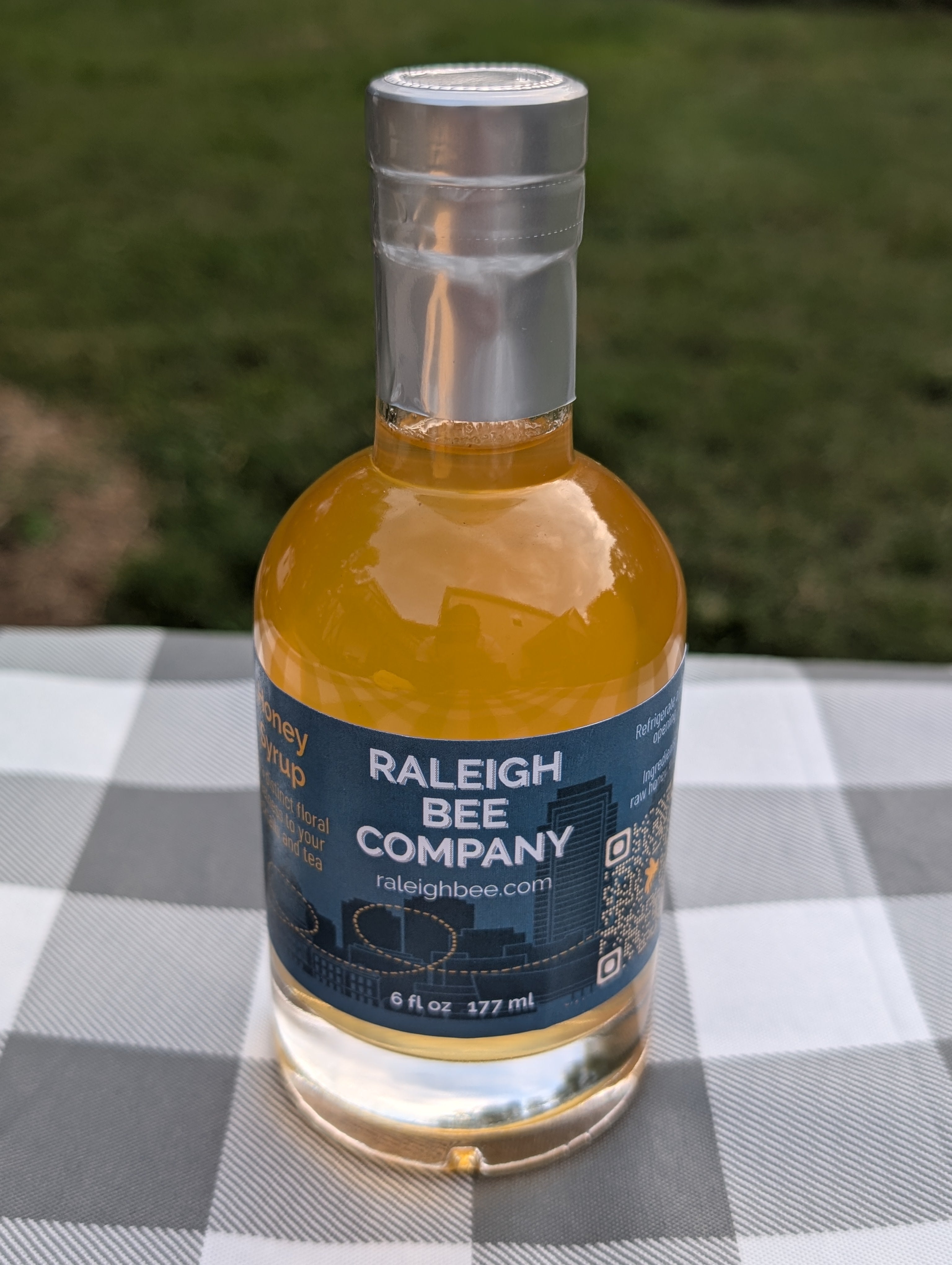 Bottle of Raleigh Bee Company honey syrup on a checkered tablecloth with a blurred green background